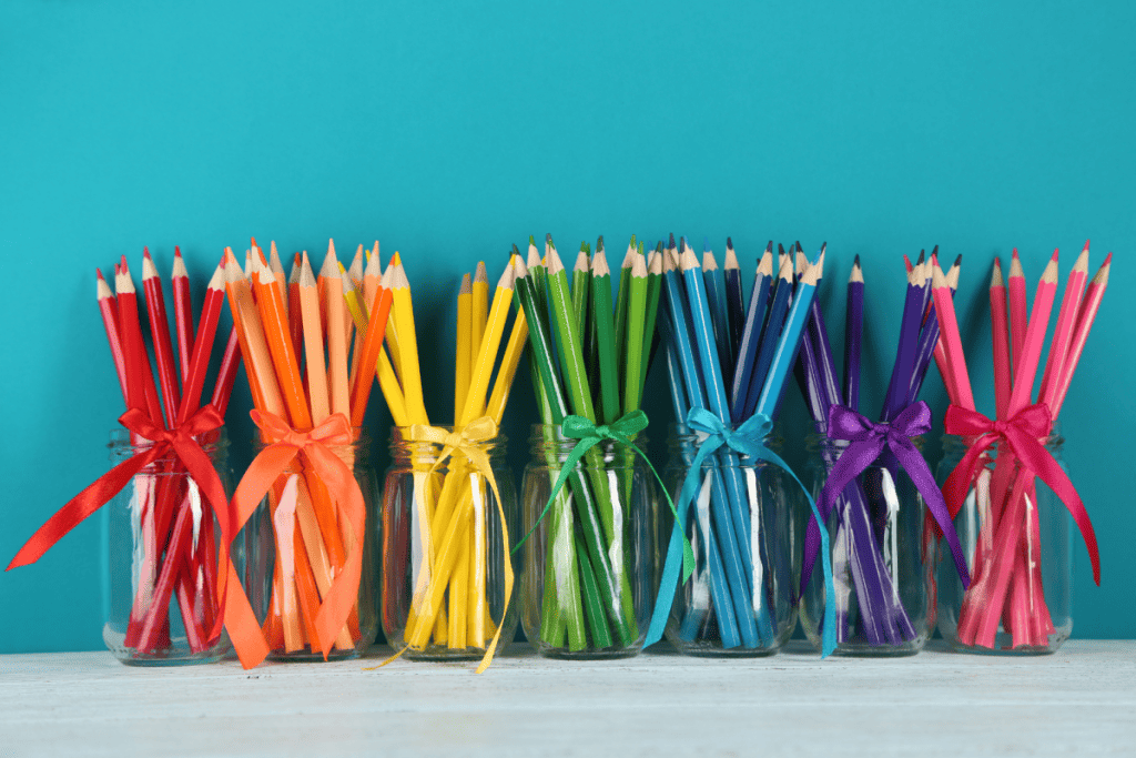 Seven glass candle containers reused as colored pencil holders. Each jar has a ribbon that corresponds to the color of the pencils inside the container.