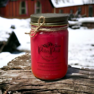 A red candle with the scent of Christmas Cabin sitting on wood. In the background there is snow on the ground and a red cabin.