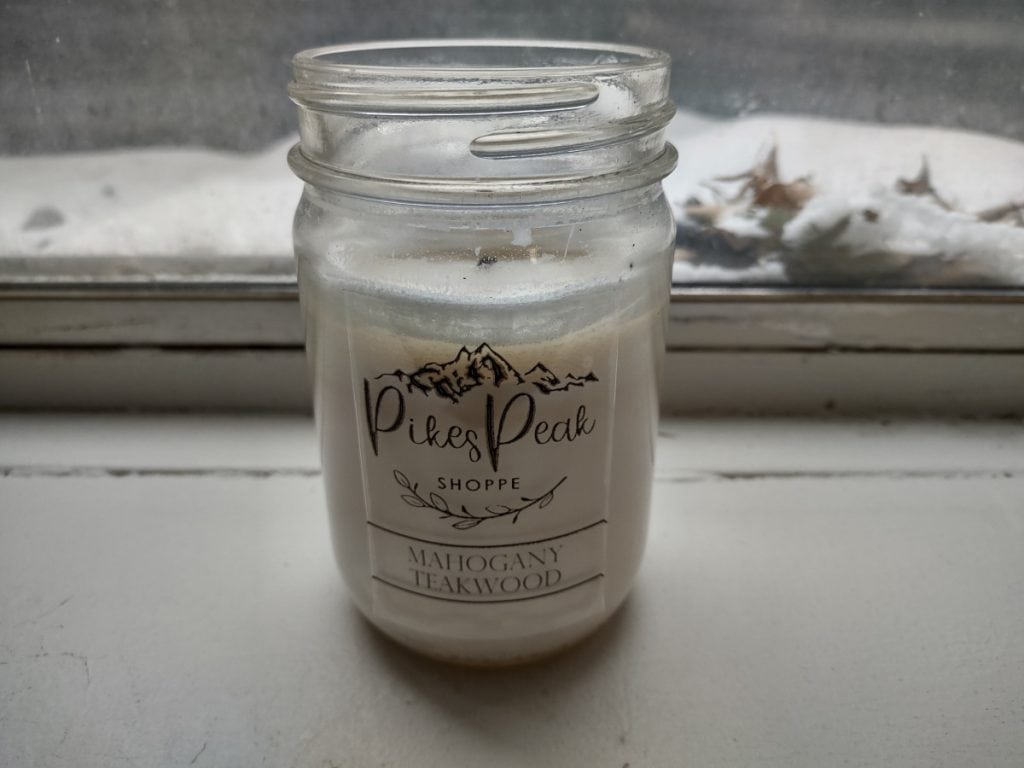 A white Mahogany Teakwood candle from Pikes Peak Shoppe on a white window sill with snow and leaves outside the window