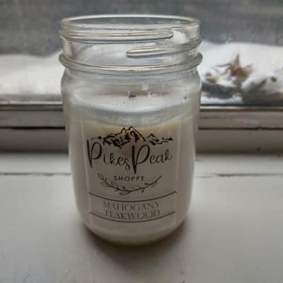 A white Mahogany Teakwood candle from Pikes Peak Shoppe on a white window sill with snow and leaves outside the window