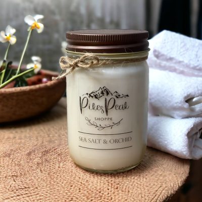 A white candle from Pikes Peak Shoppe with the scent of Sea Salt and Orchid. The candle is sitting on a peach cloth and in the background there are white folded towels and part of a flower.
