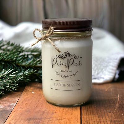 A white candle in the scent of Tis The Season. The candle is sitting on some wood and in the background there are needles from an evergreen tree and a white towel draped over something